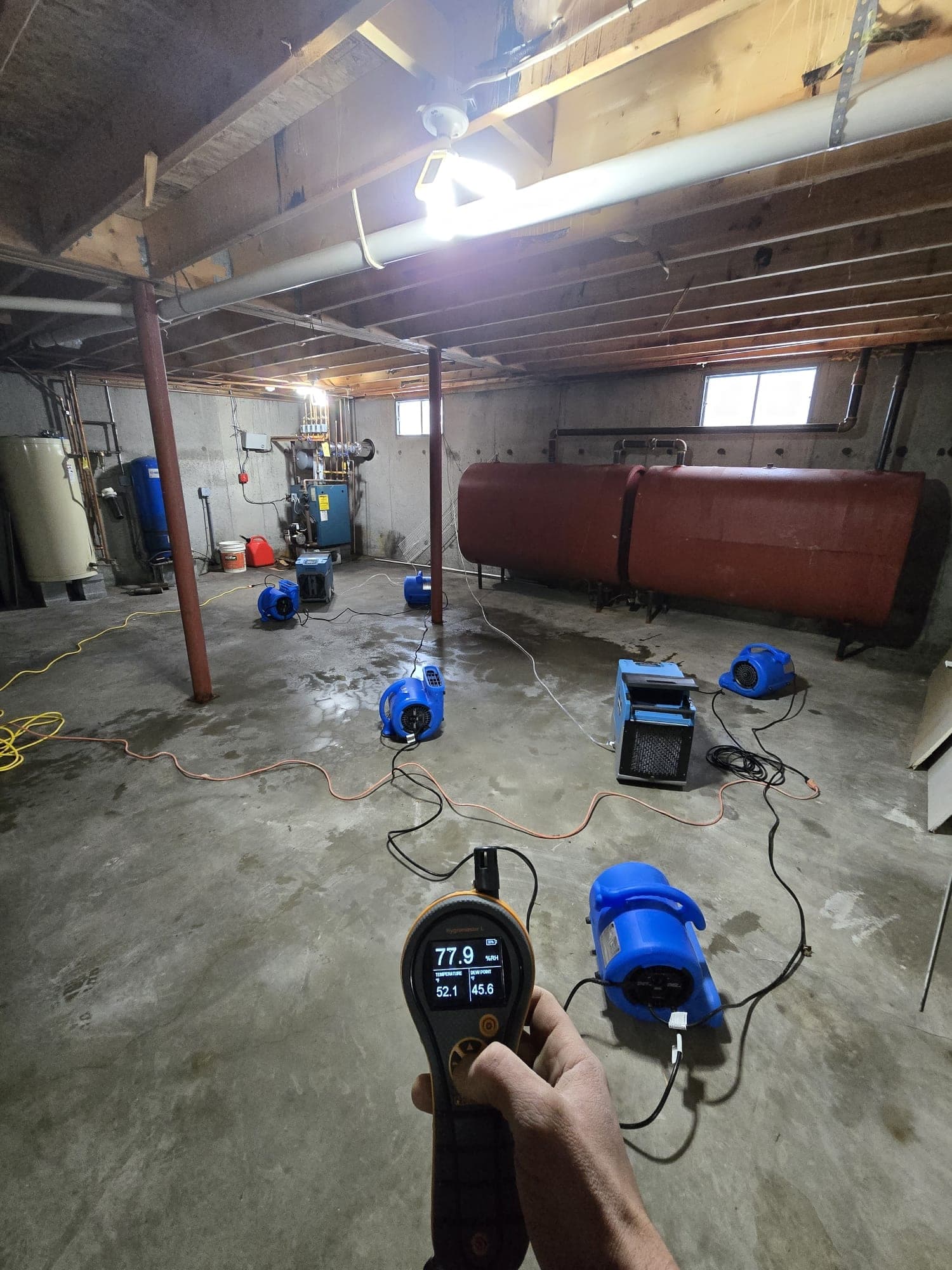 Technician checking a thermo-hygrometer while air movers and a dehumidifier dry a basement floor