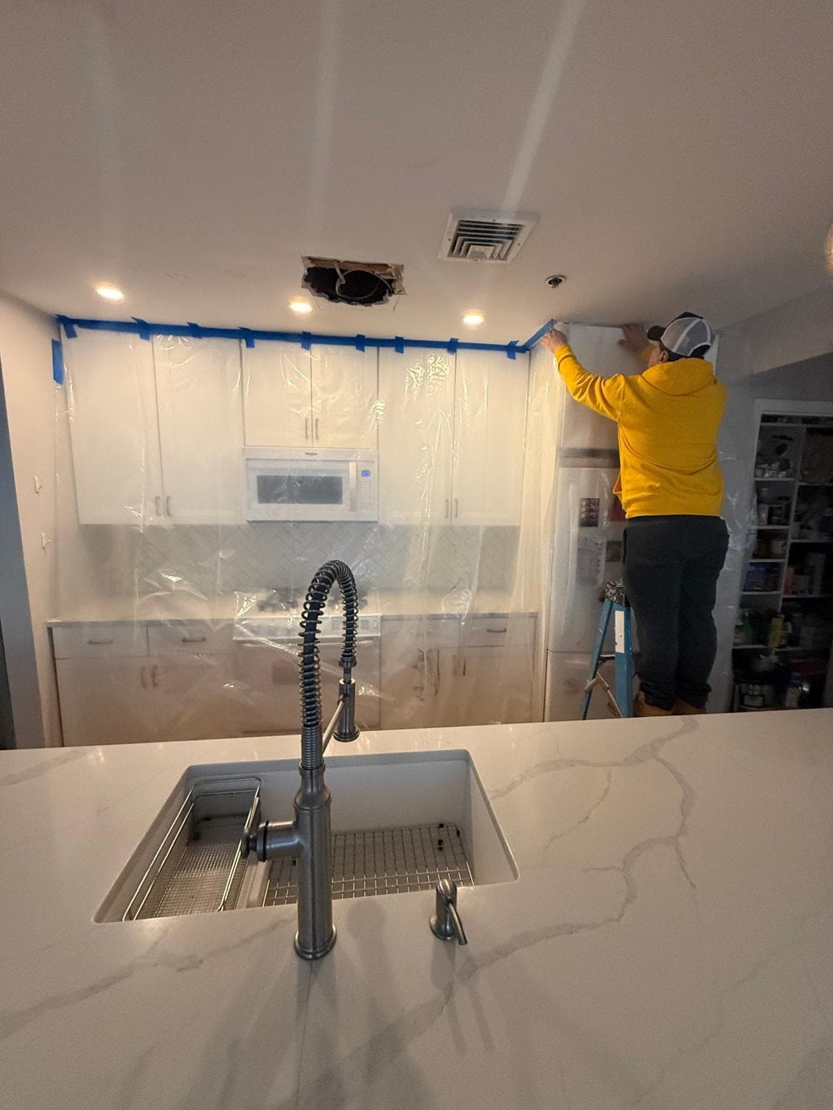 Technician on a ladder taping poly sheeting over kitchen cabinets under a water-damaged ceiling