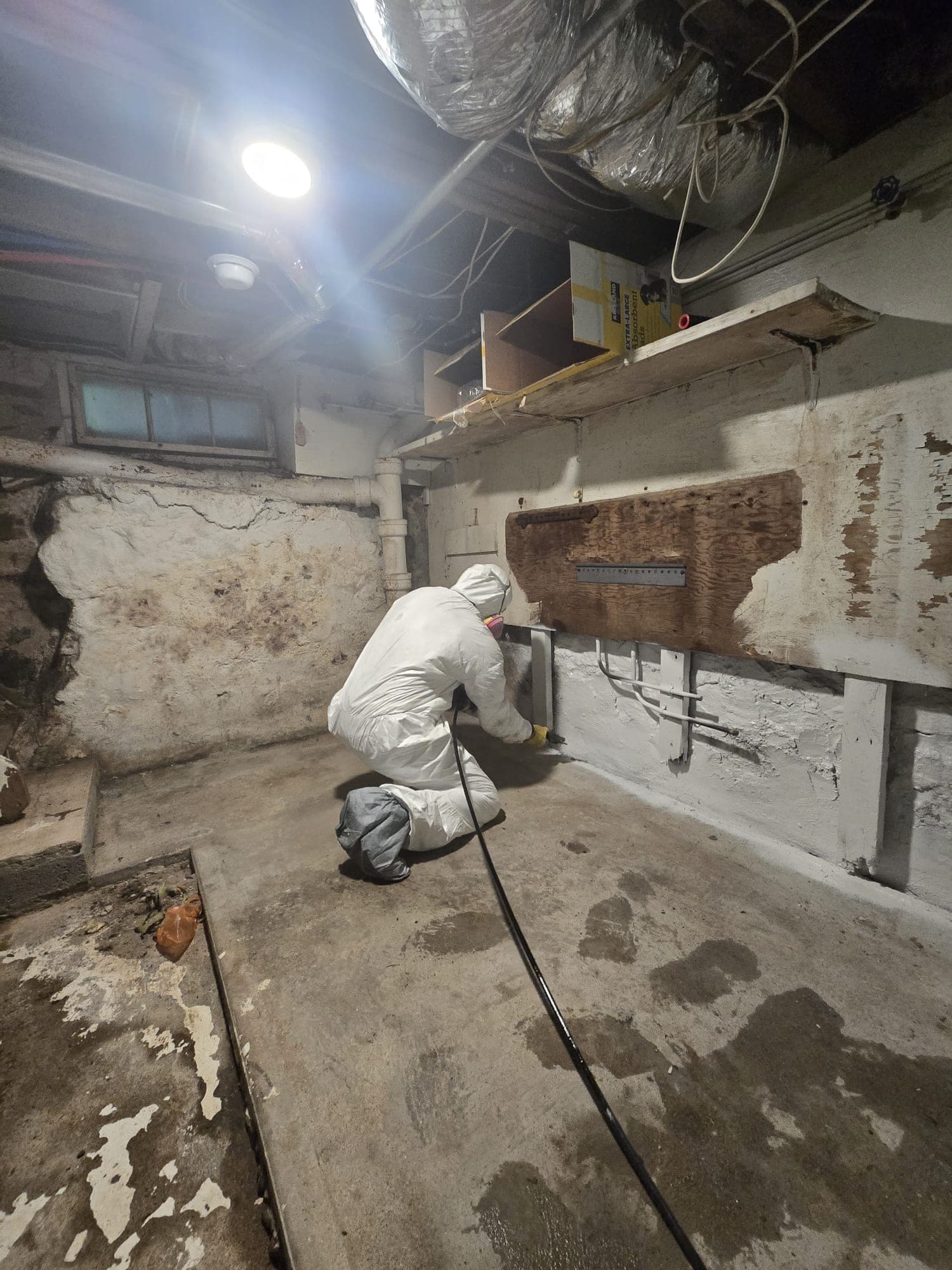 Technician in a protective suit applying sealant along the base of a stone foundation wall