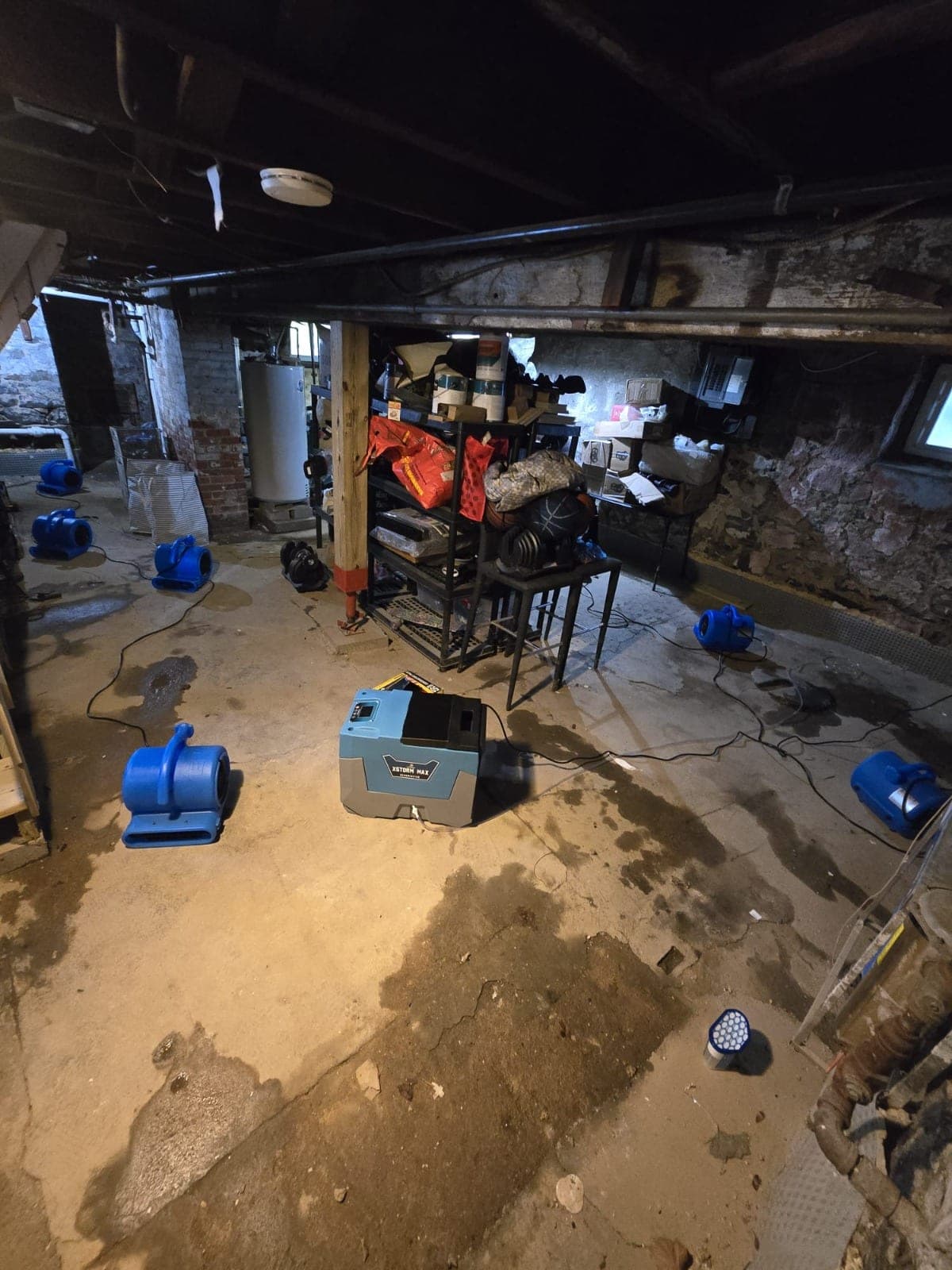 Air movers and a dehumidifier drying a damp concrete floor in a stone-foundation basement