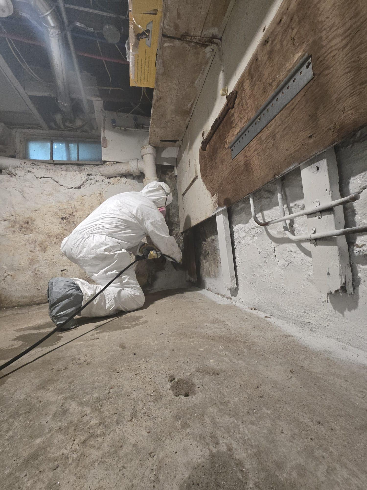 Technician in a protective suit sealing a foundation wall around supply pipes after mold removal